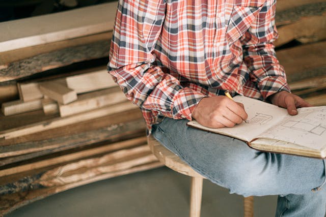 Man in a plaid shirt sketching in front of a pile of rough cut lumber.